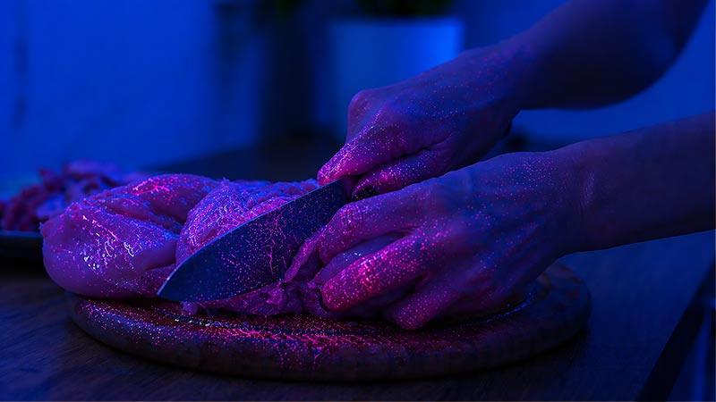 Hands cutting raw chicken under UV light showing contamination concentrated on fingers and meat