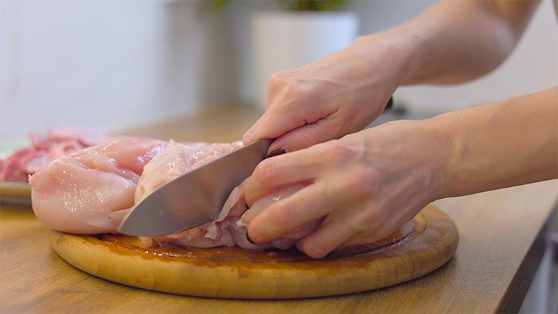 Hands cutting raw chicken on a chopping board in normal lighting